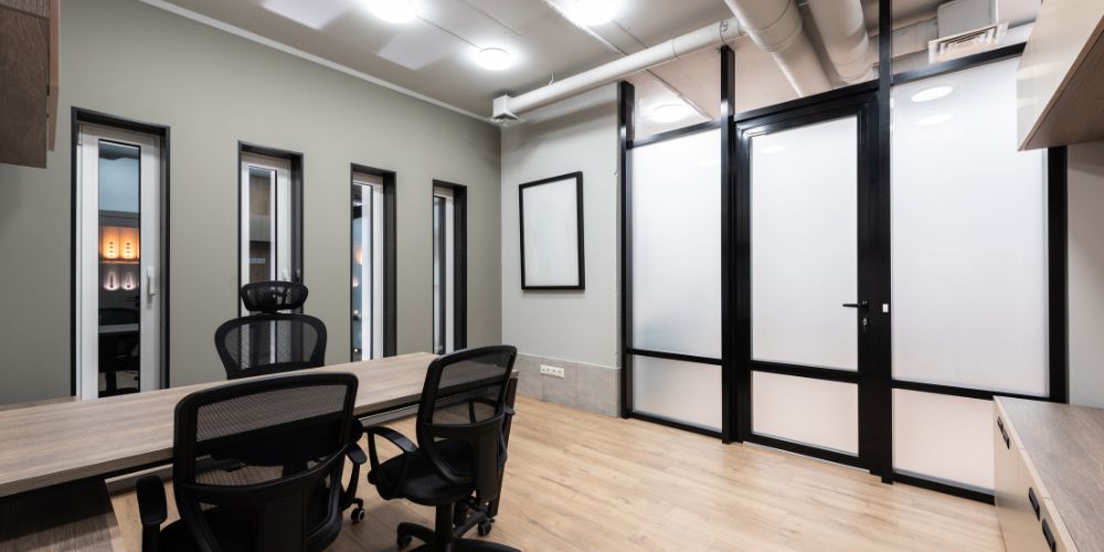 Modern office interior with glass partitions, wooden flooring, ergonomic chairs, and overhead lighting.